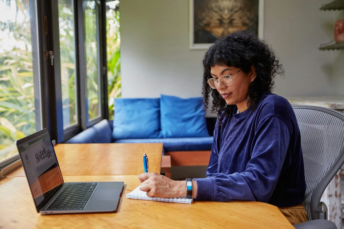 A woman writing notes at a table at home with a laptop open beside her